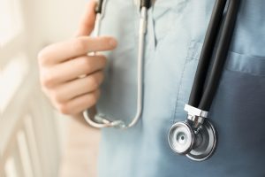 A young female doctor in a blue smock holding a phonendoscope in arms. Blurred background. Concept of medical help. Light effect.